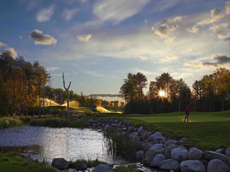 Golf course green at sunset with Toro irrigation sprinklers running beside a water feature, showcasing sustainable turf management.